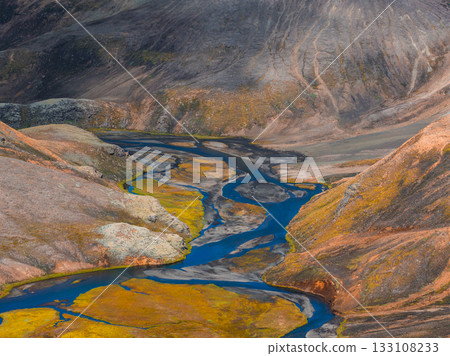 A vibrant blue river winds through a rugged Icelandic terrain of orange, brown, and yellow hues, with green moss and rolling hills in the background. A vibrant blue river winds through a rugged Icelandic terrain of orange, brown, and yellow hues, with green moss and rolling hills in the background. 133108233