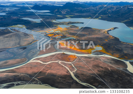Aerial image of braided rivers flowing through volcanic terrain with orange and yellow hues, surrounded by lakes and rugged mountains in Iceland. Aerial image of braided rivers flowing through volcanic terrain with orange and yellow hues, surrounded by lakes and rugged mountains in Iceland. 133108235