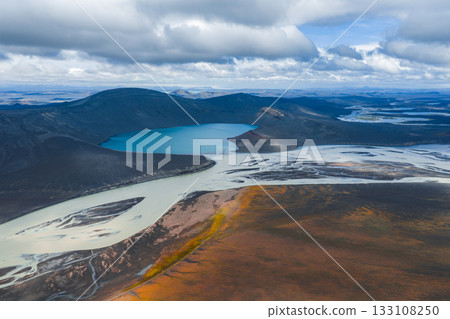 Aerial image of a vibrant blue lake with braided rivers, surrounded by dark volcanic hills, orange and yellow vegetation, and a cloudy sky in Iceland. Aerial image of a vibrant blue lake with braided rivers, surrounded by dark volcanic hills, orange and yellow vegetation, and a cloudy sky in Iceland. 133108250