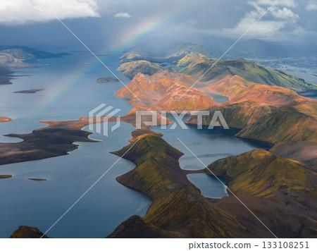 Aerial view of Iceland's rhyolite mountains with vivid colors, interconnected lakes, and a rainbow under a partly cloudy sky in the Landmannalaugar region. 133108251