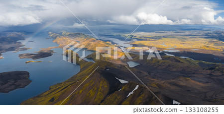 Aerial view of Iceland's highlands with vibrant hills, deep blue lakes, volcanic rock, green moss, snow patches, and a faint rainbow under cloudy skies. 133108255