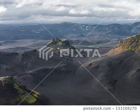 Dark volcanic terrain with vibrant green moss, rugged hills, and distant mountains under a cloudy sky, creating a dramatic Icelandic scene. 133108270