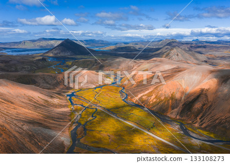 Aerial view of Iceland's colorful highlands with orange rhyolite hills, green mossy valleys, winding rivers, distant mountains, and a lake under a blue sky. 133108273