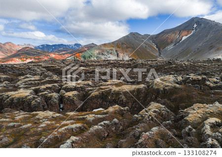 Volcanic lava fields in the foreground with colorful rhyolite mountains in the background, featuring orange, green, and gray hues under a partly cloudy sky. 133108274