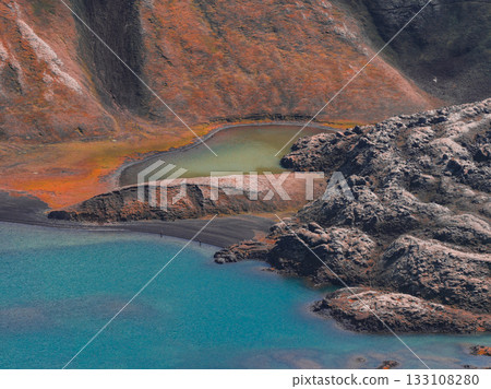 A turquoise lake bordered by rocky terrain, a smaller greenish yellow lake, and steep hills with orange and brown hues in Iceland's landscape. 133108280