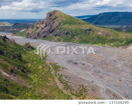 Aerial view of a green covered mountain in Iceland with steep rocky slopes, braided river channels on a gravel plain, and distant hills under a partly cloudy sky. Aerial view of a green covered mountain in Iceland with steep rocky slopes, braided river channels on a gravel plain, and distant hills under a partly cloudy sky. 133108290