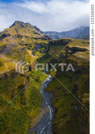 Mulagljufur Canyon in Iceland features steep moss covered cliffs, a winding river, vibrant green vegetation, and distant snow capped peaks under clouds. Mulagljufur Canyon in Iceland features steep moss covered cliffs, a winding river, vibrant green vegetation, and distant snow capped peaks under clouds. 133108303