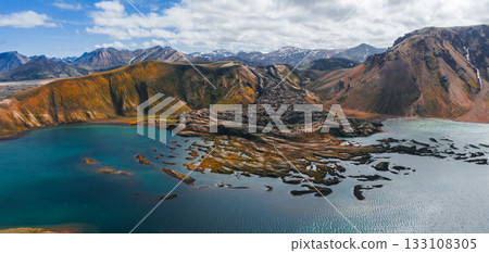 Aerial view of vibrant Icelandic mountains with orange, green, and brown hues, a lava field, a serene lake, and snow capped peaks in the background. 133108305