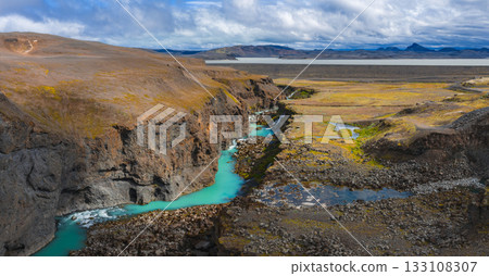 Turquoise river flows through Sigoldugljufur Canyon in Iceland, surrounded by rocky cliffs, mossy terrain, scattered pools, and distant plains. 133108307