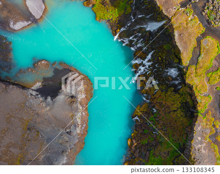 Turquoise river winds through volcanic rock and green moss in Iceland. Small waterfalls cascade along the river, adding movement to the rugged terrain. 133108345