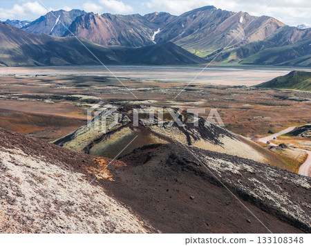 Multicolored rhyolite mountains, a volcanic hill with moss and lava rock, and a dirt road with a small vehicle in a remote Icelandic valley. 133108348