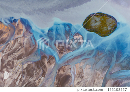 Vibrant blue glacial streams flow through brown terrain, surrounding a moss covered island in Iceland. The intricate patterns create striking contrasts. 133108357