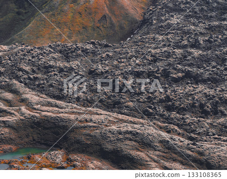Rugged volcanic terrain in Iceland with dark lava fields, earthy orange and green slopes, and a vivid green and orange pool in the foreground. 133108365