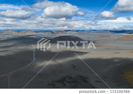 A barren volcanic terrain in Iceland with ash covered ground, red craters, a winding dirt road, and a cloudy sky casting dynamic shadows. 133108366