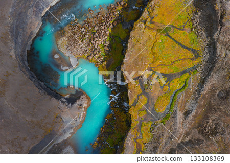 Turquoise river winds through rocky terrain and moss covered ground in Iceland. Aerial perspective highlights vivid colors and natural patterns. 133108369