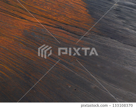 Aerial view of a barren volcanic terrain in Iceland, featuring dark ash covered ground streaked with vibrant orange and rust colored patterns. 133108370