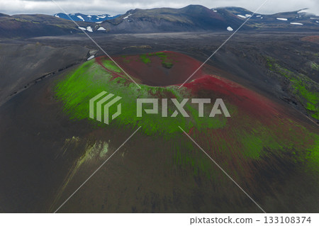 Aerial view of a volcanic crater in Iceland with green moss, red minerals, dark lava fields, and distant snow capped mountains under a cloudy sky. 133108374