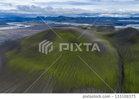 Aerial view of green moss covered hills on dark volcanic soil, braided rivers on a plain, and distant mountains under a partly cloudy sky in Iceland. 133108375