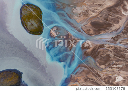 Aerial image of a braided glacial river in Iceland with vibrant blue water, rugged brown terrain, and two moss covered islands as focal points. 133108376