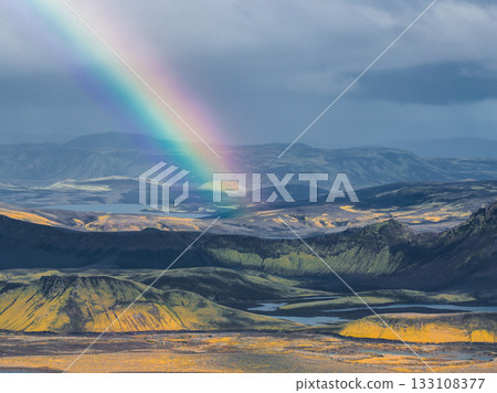Aerial view of Icelandic highlands featuring a rainbow, rolling hills, volcanic craters, mossy vegetation, dark rocks, small lakes, and streams. 133108377
