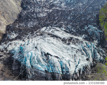 A glacier in Iceland with streaks of white and blue ice, dark volcanic ash, steep rocky cliffs, and green vegetation creating a dramatic contrast. 133108380