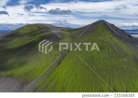 Two green moss covered volcanic hills in Iceland with streaks of vegetation and dark soil, set against a blue sky with scattered clouds. Two green moss covered volcanic hills in Iceland with streaks of vegetation and dark soil, set against a blue sky with scattered clouds. 133108385