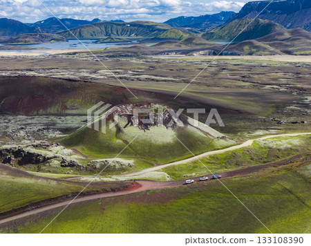 A volcanic crater with a dark rocky rim surrounded by moss covered terrain in Iceland. A dirt road and vehicles highlight the scale, with hills and water in the background. 133108390
