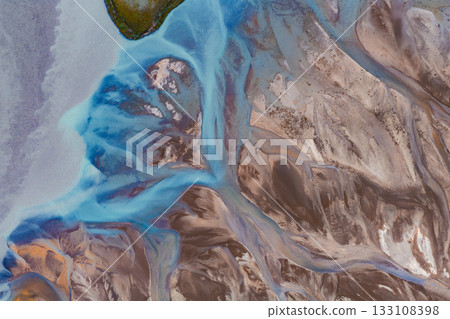 Braided river system with vibrant blue glacial waters flowing through earthy tones, featuring a small green island and intricate geological patterns. 133108398