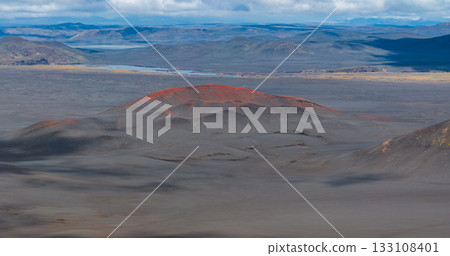 A red hued volcanic crater contrasts with dark ash covered terrain in Iceland. Rolling hills, distant mountains, and a faint river complete the scene. A red hued volcanic crater contrasts with dark ash covered terrain in Iceland. Rolling hills, distant mountains, and a faint river complete the scene. 133108401