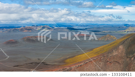 A barren volcanic terrain in Iceland with red and orange craters, a winding dirt road, yellow moss covered slopes, distant mountains, and a blue sky. 133108411