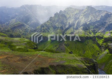 Aerial view of moss covered peaks and steep cliffs in Mulagljufur Canyon, Iceland. Light mist drifts over the rugged landscape, adding texture and depth. 133108413