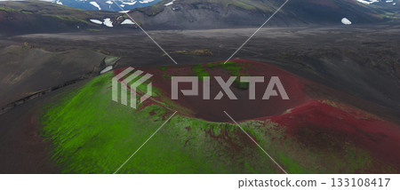 A volcanic crater in Iceland with red soil and green moss, surrounded by a dark volcanic plain. Snow capped mountains are visible in the distance. A volcanic crater in Iceland with red soil and green moss, surrounded by a dark volcanic plain. Snow capped mountains are visible in the distance. 133108417