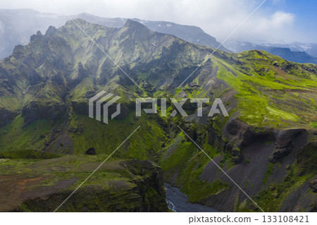 Lush green canyon in Iceland with moss covered cliffs, a winding river, and misty mountain peaks partially obscured by clouds. 133108421