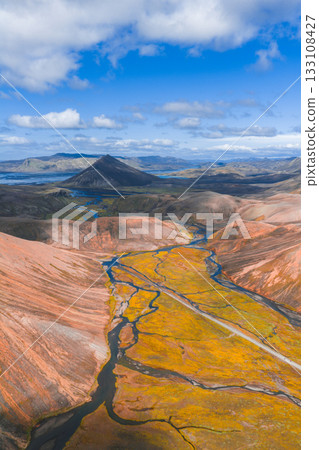 Aerial view of Iceland's Landmannalaugar region with colorful rhyolite mountains, a winding river, golden moss, and a conical mountain under a blue sky. 133108427