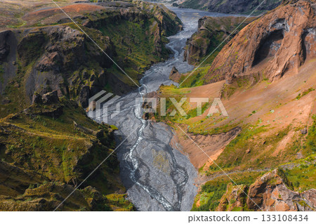 Aerial perspective of Mulagljufur Canyon in Iceland, featuring dramatic cliffs, vibrant green vegetation, a winding river, and a prominent arched rock face. 133108434