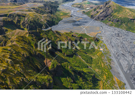 Aerial view of a rugged Icelandic valley with a winding riverbed, steep green cliffs, volcanic rock, and rolling hills under natural light. 133108442