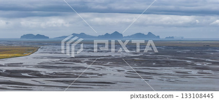 Aerial view of braided river patterns on a dark plain with Vestmannaeyjar islands in the distance, set beneath a cloudy and dramatic sky. Aerial view of braided river patterns on a dark plain with Vestmannaeyjar islands in the distance, set beneath a cloudy and dramatic sky. 133108445