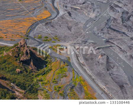 Aerial view of a braided river system in Iceland with a small bridge, rocky outcrop, green vegetation, autumn hues, and vehicles near the outcrop. 133108446