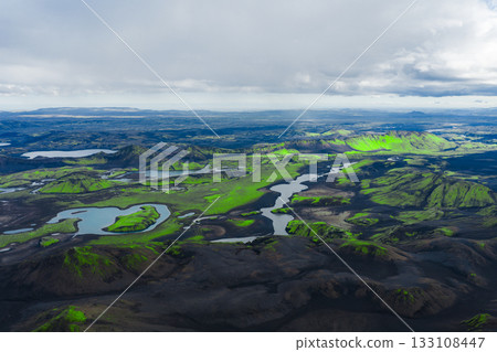 Aerial view of green moss covered hills, dark volcanic terrain, winding rivers, and small lakes under a partly cloudy sky in Iceland. 133108447