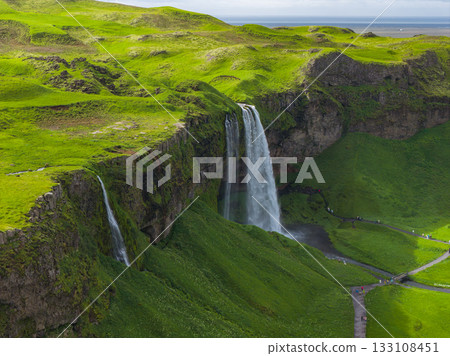 Seljalandsfoss waterfall cascades from a tall cliff in Iceland, surrounded by green moss and grass. A path and bridge lead close, with the ocean in view. 133108451