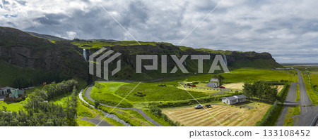 Lush green fields, scattered farmhouses, and cascading waterfalls on a dramatic cliffside in Iceland, with a winding road and stream in the valley. 133108452