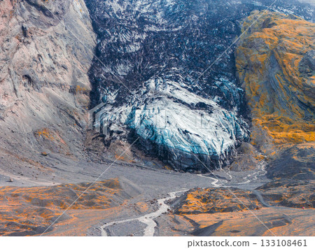 A glacier with icy blue and white surface lies between dark volcanic cliffs. A winding stream flows through gray and orange terrain in Iceland. 133108461
