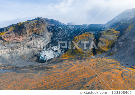 A dramatic Icelandic canyon with steep, colorful rock formations and a glacier nestled between cliffs under a cloudy sky, creating a striking scene. 133108465
