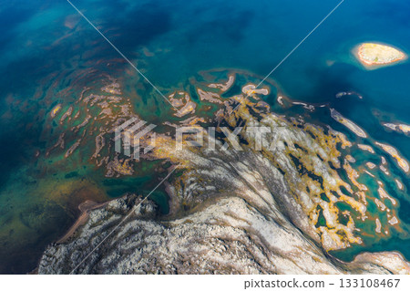 Aerial view of deep blue waters with golden and green patterns of algae or minerals, featuring a small island in the upper right corner in Iceland. Aerial view of deep blue waters with golden and green patterns of algae or minerals, featuring a small island in the upper right corner in Iceland. 133108467