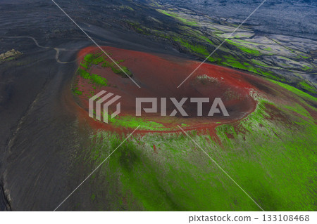 Aerial view of a volcanic crater in Iceland with red volcanic soil, green moss covered slopes, and a barren surrounding landscape with a winding road. Aerial view of a volcanic crater in Iceland with red volcanic soil, green moss covered slopes, and a barren surrounding landscape with a winding road. 133108468