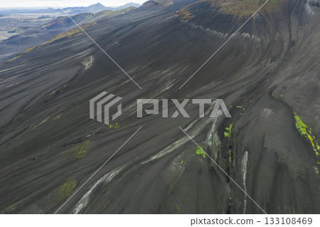 Dramatic aerial view of Iceland's volcanic terrain with black ash slopes, vibrant green moss patches, and distant mountains in the background. Dramatic aerial view of Iceland's volcanic terrain with black ash slopes, vibrant green moss patches, and distant mountains in the background. 133108469