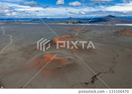 Aerial view of red and orange volcanic craters in Iceland, surrounded by dark soil, a winding dirt road, distant mountains, and a lake under clouds. Aerial view of red and orange volcanic craters in Iceland, surrounded by dark soil, a winding dirt road, distant mountains, and a lake under clouds. 133108470