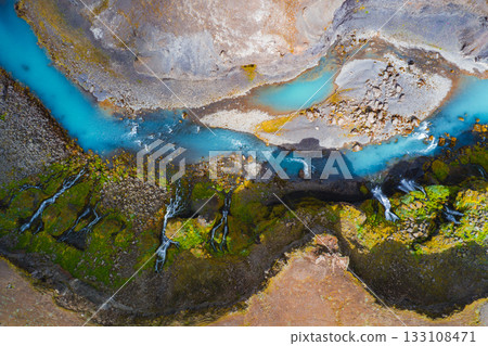 Turquoise river winds through moss covered cliffs with cascading waterfalls in Iceland. Contrasting colors of water, vegetation, and terrain are visible. 133108471