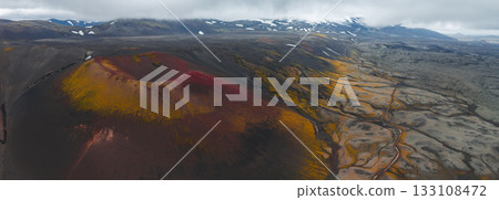 Aerial view of a red and yellow crater in Iceland's volcanic terrain, with snow capped mountains, a winding road, and low hanging clouds in the background. 133108472