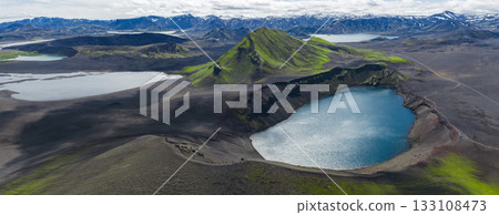 Aerial view of a vivid blue crater lake surrounded by dark volcanic terrain, a green moss covered mountain, and snow capped peaks in Iceland. Aerial view of a vivid blue crater lake surrounded by dark volcanic terrain, a green moss covered mountain, and snow capped peaks in Iceland. 133108473